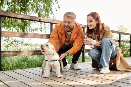 A couple in casual attire pet a dog on a wooden deck in the park.