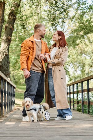 A man and woman in casual attire stand on a bridge with their dog in a park.