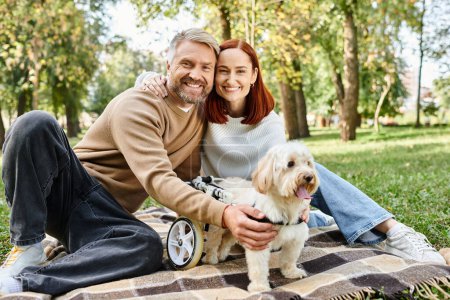 A loving couple and their dog sit on a checkered blanket in a park.