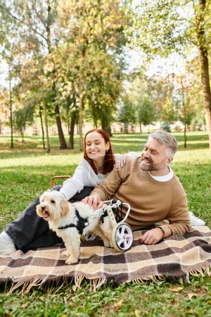 Un hombre y una mujer en traje casual se sientan en una manta con su perro en un entorno tranquilo parque.
