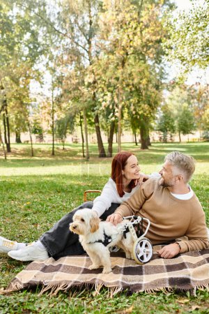 A man and woman sit on a blanket with their dog in a park.