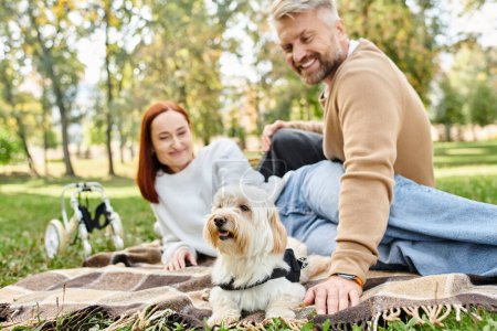 An adult loving couple sits on a blanket with their dog in a beautiful park setting.