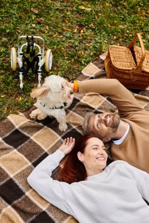 A man and woman relax on a blanket with their dog in a park.