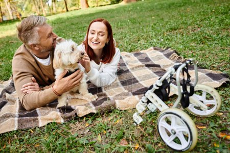 A man and woman lay on a blanket with their dog in a park.