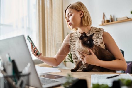 Une femme aux cheveux courts tient un chat tout en se concentrant sur un écran d'ordinateur portable à la maison.