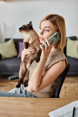 Eine Frau führt interaktiv ein Telefongespräch, während sie ihre Katze liebevoll an einem Tisch hält.
