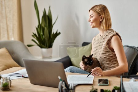 Une femme aux cheveux courts s'assoit à une table avec un ordinateur portable, tapant, tandis que son chat regarde avec curiosité.