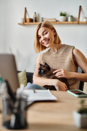 A relaxed woman with short hair seated at a desk, holding a cat in her arms, sharing a peaceful moment at home.