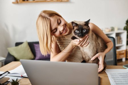 Foto de Una mujer con el pelo corto sostiene a su gato mientras mira la pantalla de un ordenador portátil en un ambiente acogedor en casa. - Imagen libre de derechos