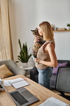 Foto de Una mujer serena con el pelo corto sostiene a su gato, de pie ante un ordenador portátil en un ambiente acogedor en casa. - Imagen libre de derechos