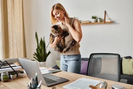 Foto de Una mujer sostiene a su gato amorosamente mientras está de pie junto a una computadora portátil en casa. - Imagen libre de derechos