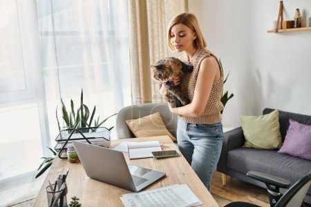 Una mujer con el pelo corto de pie delante de un ordenador portátil, sosteniendo a su gato en un momento acogedor en casa.