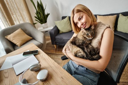 Foto de Una mujer con el pelo corto sentado en una mesa, sosteniendo y acariciando a su gato en un momento tranquilo en casa. - Imagen libre de derechos