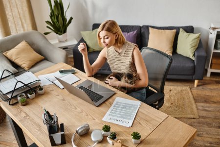 Téléchargez les photos : Une femme élégante aux cheveux courts assise à une table avec un ordinateur portable, accompagnée de son chat dans un cadre confortable à la maison. - en image libre de droit