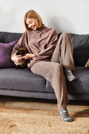 A woman with short hair relaxing on a couch, holding and petting her cat with care and love.