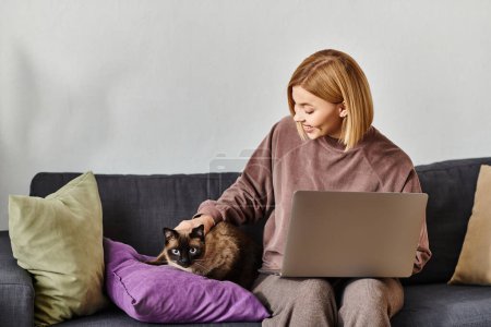 A woman with short hair sits on a couch with a laptop, petting her cat.