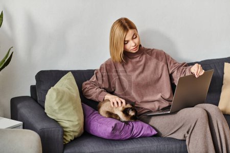 Une femme sereine aux cheveux courts se relaxant sur un canapé avec un chat paisible sur ses genoux à la maison.