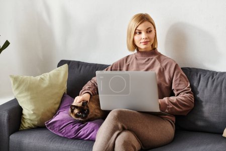 Une femme aux cheveux courts s'assoit sur un canapé, à l'aide d'un ordinateur portable, à côté de son chat.