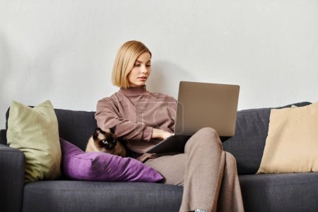 Eine stylische Frau mit kurzen Haaren, die sich auf ihren Laptop konzentriert, während sie es sich auf einer bequemen Couch bequem macht, mit ihrer Katze an ihrer Seite.