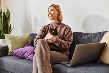 Une femme aux cheveux courts assise sur un canapé, berçant un chat dans ses bras, à la fois content et paisible.