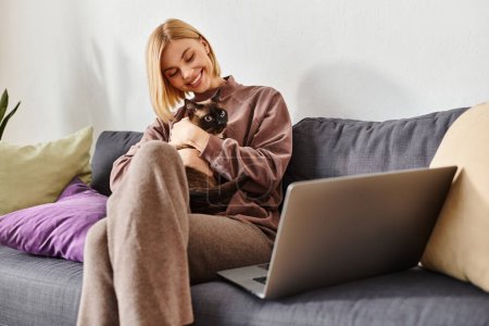 Une femme sereine aux cheveux courts assise sur un canapé, tenant un chat dans un moment paisible à la maison.