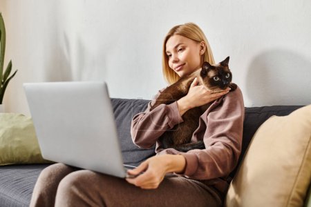 Eine Frau mit kurzen Haaren genießt die Zeit zu Hause, hält eine Katze, während sie einen Laptop auf einer gemütlichen Couch benutzt.