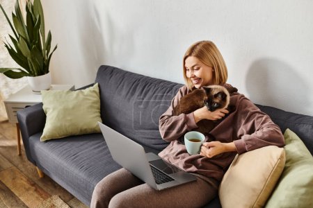 Una mujer con el pelo corto sentado en un sofá, abrazando a un gato y trabajando en un ordenador portátil en un ambiente acogedor en casa.