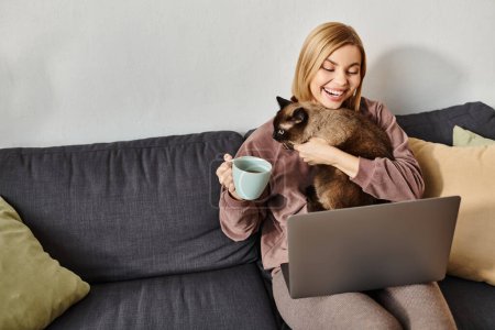 Une femme aux cheveux courts assise sur un canapé, dégustant une tasse de café tout en tenant son chat dans ses bras.