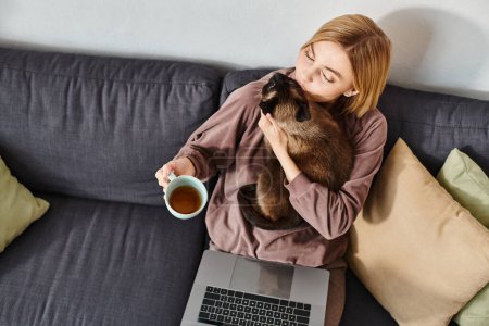 Eine schicke Frau entspannt sich auf einem Sofa und wiegt ihre pelzige Katze mit einem zufriedenen Blick in den Arm..