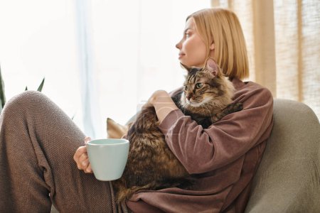 Una mujer serena con el pelo corto se sienta en un sofá sosteniendo una taza de café y un gato amistoso en un ambiente acogedor en casa.