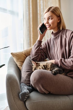 Eine stylische Frau mit kurzen Haaren plaudert auf ihrem Handy und teilt einen Moment mit ihrer entzückenden Katze auf der Couch.