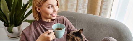 A woman with short hair sits on a couch, holding a cup of coffee and a cat in her arms, enjoying a cozy moment at home.