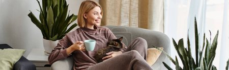 Una mujer con el pelo corto se sienta en una silla, sosteniendo una taza mientras su gato curiosamente se acurruca cerca.