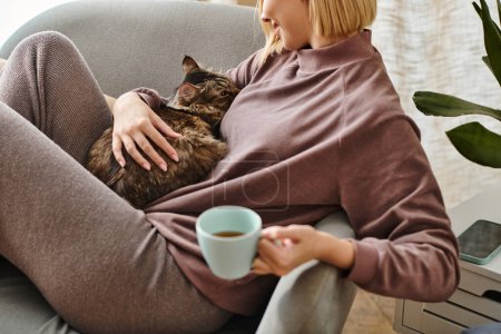 Eine Frau mit kurzen Haaren entspannt sich auf einer Couch, wiegt eine Tasse Kaffee und eine zufriedene Katze in ihren Armen.