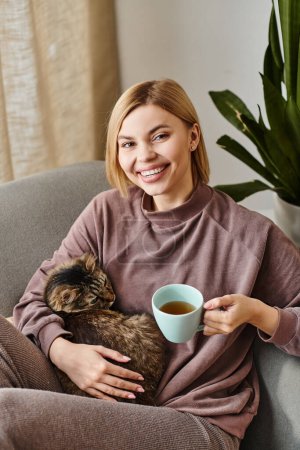 A woman with short hair relaxes on a couch, cradling a cup of coffee while her cat snuggles in her lap.