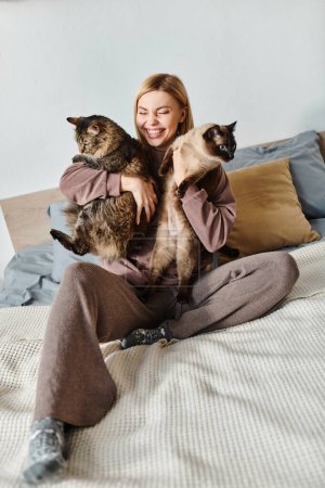 A serene woman with short hair sits on a bed, holding two cats close to her, enjoying a peaceful moment at home.