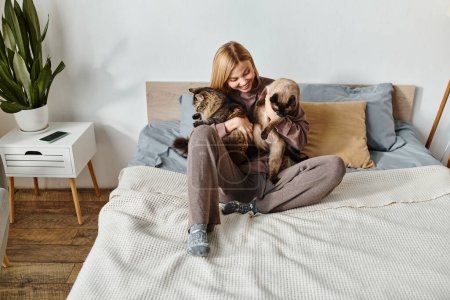 Une femme aux cheveux courts assise sur un lit, tenant paisiblement deux chats dans ses bras.