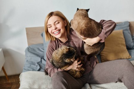 Eine Frau mit kurzen Haaren sitzt auf einem Bett, hält zärtlich zwei Katzen in ihren Armen und genießt einen friedlichen Moment zu Hause.