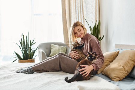 Eine Frau mit kurzen Haaren sitzt auf einem Bett und hält sanft eine Katze in ihren Armen, während sie einen ruhigen und friedlichen Moment miteinander teilt..