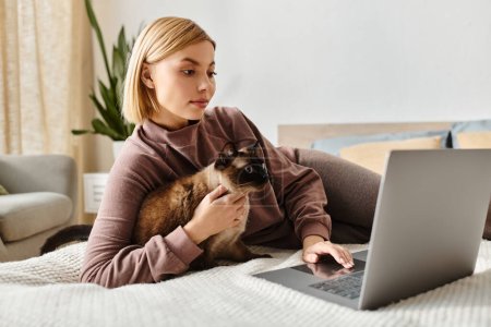 A stylish woman with short hair sits on a bed, typing on a laptop while her fluffy cat rests beside her.