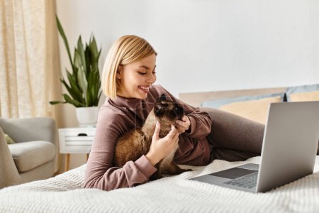 Short-haired woman relaxing on a bed with her cat while using a laptop.