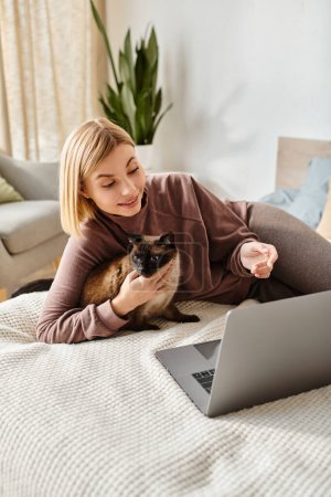 Una mujer con el pelo corto se relaja en una cama con su gato, absorto en su ordenador portátil.
