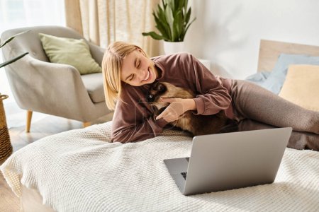 A stylish woman with short hair relaxes on a bed, engrossed in her laptop while her cat cuddles beside her.