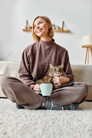 Photo for A woman with short hair peacefully sits on the floor, cradling her cat in her arms at home. - Royalty Free Image