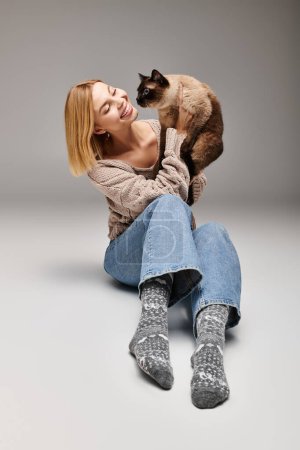 A stylish woman with short hair sits on the floor, her feline companion perched on her shoulders.