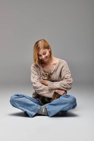 Eine Frau mit kurzen Haaren sitzt auf dem Boden und hält ihre Katze in liebevoller Umarmung in einer gemütlichen häuslichen Umgebung.