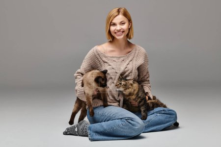 Eine Frau mit kurzen Haaren sitzt auf dem Boden, umgeben von zwei Katzen, und genießt einen friedlichen Moment zusammen zu Hause.