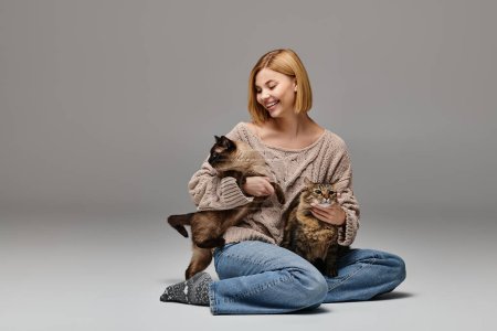 Une femme aux cheveux courts assise par terre, tenant amoureusement deux chats dans un moment serein et paisible à la maison.