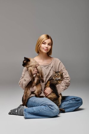 Une femme aux cheveux courts s'assoit sur le sol, berçant deux chats dans ses bras, incarnant un moment paisible et heureux à la maison.