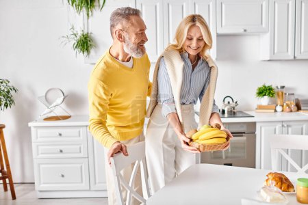 Téléchargez les photos : Un homme et une femme mûrs se tiennent dans leur cuisine confortable, tenant des bananes et partageant un moment doux ensemble. - en image libre de droit